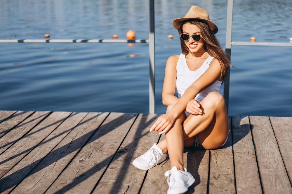 Woman wearing hat and sunglasses sits on a wooden dock by the water.