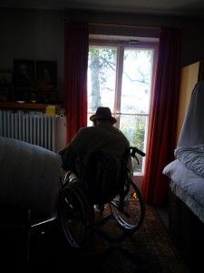 alter Mann von hinten mit Hut, sitzt in einer Wohnung im Rollstuhl
