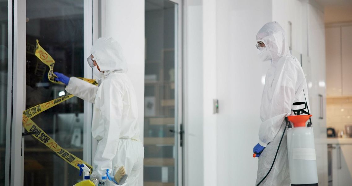 Two people in protective suits at a scene; one tapes, the other holds a sprayer.