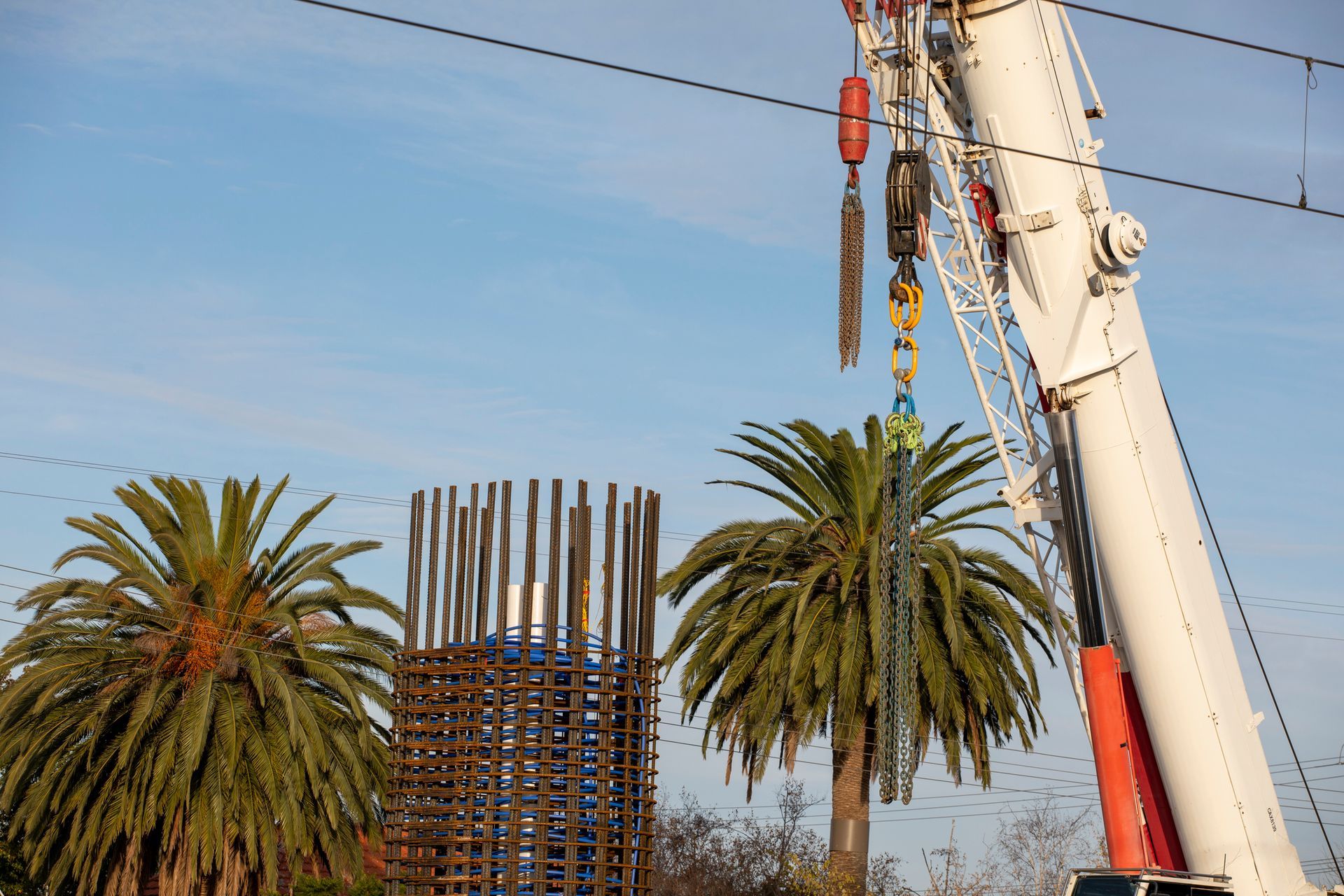 Reinforcing bars of a bridge footing under construction, with a crane boom and two palm trees | Kato