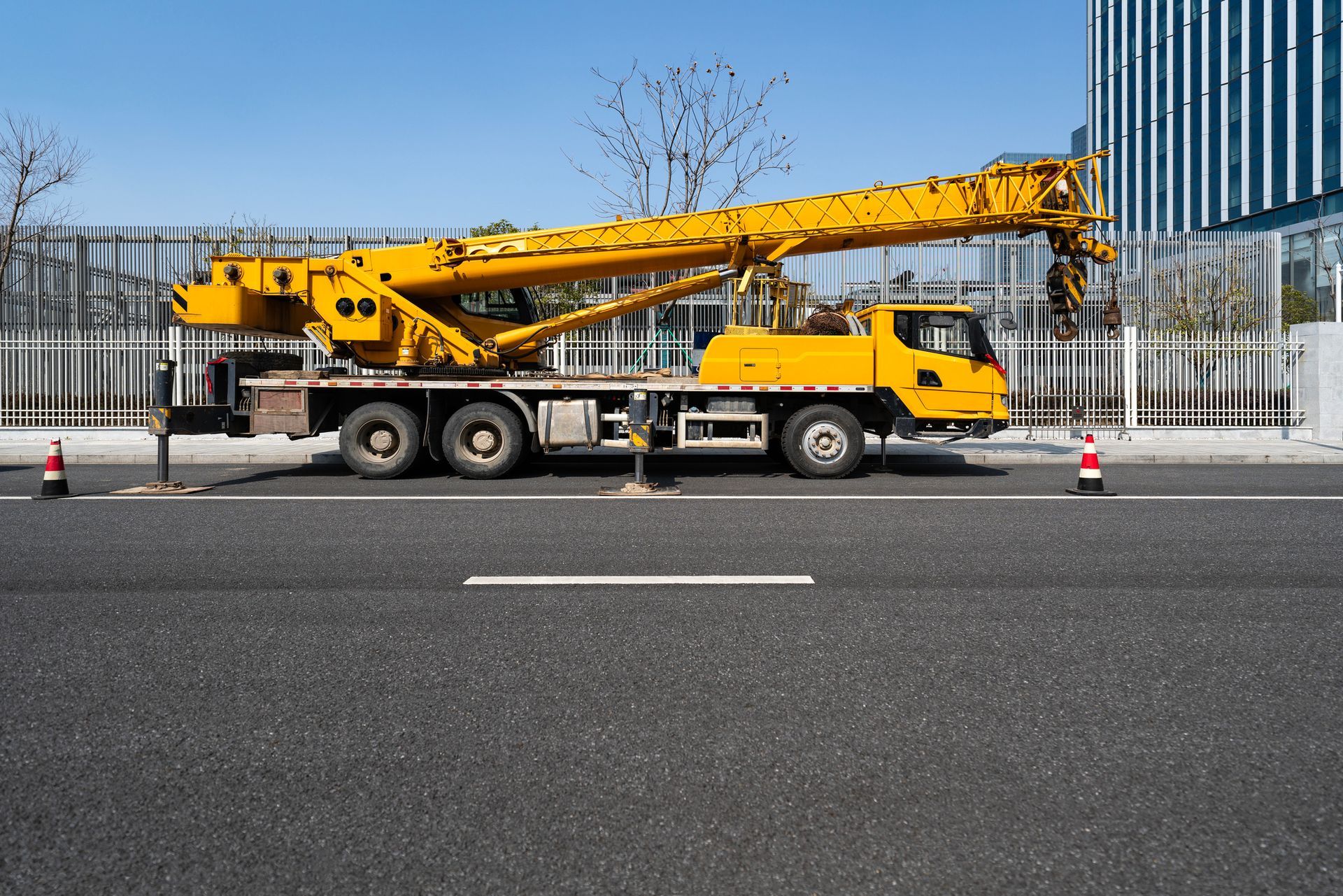 Yellow crane truck parked roadside near glass building and leafless trees.