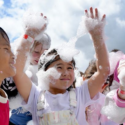 Children playing in foam, arms raised, smiling; outdoors on a sunny day.