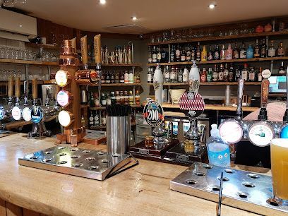Bar with beer taps, bottles of alcohol on shelves, a copper tap, and a light-colored wooden bar.