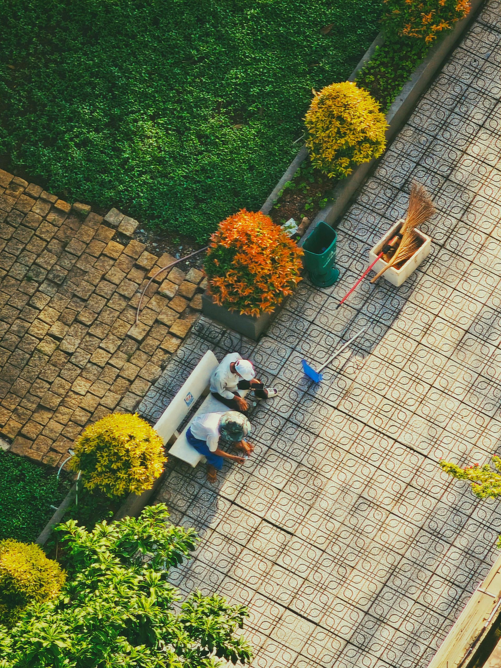 Two people sit on a park bench next to a green bin, a broom, and orange-flowering bushes on a patterned stone path.