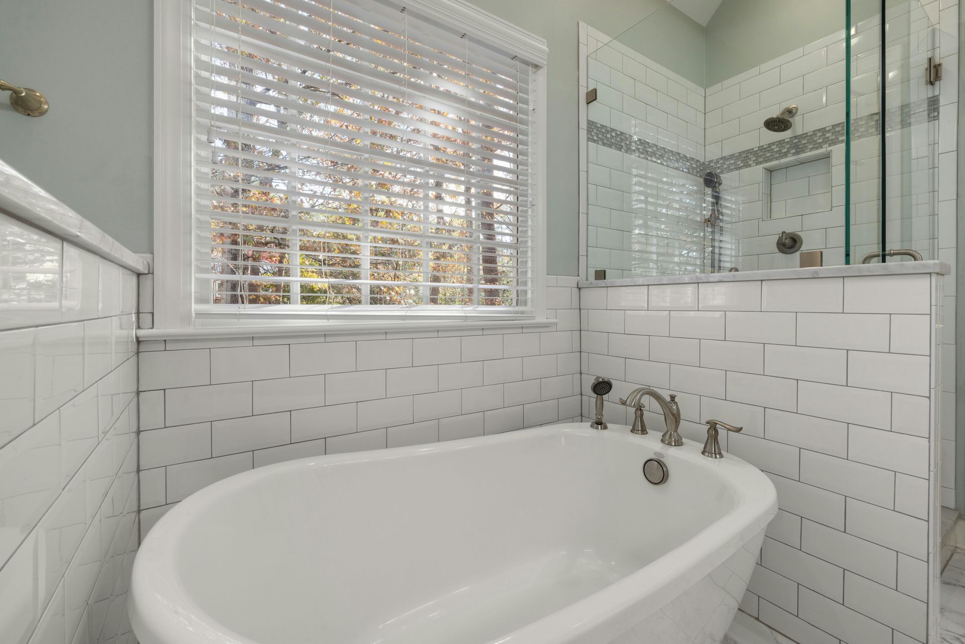 A white freestanding bathtub set against a white subway tile wall with a window and a glass shower enclosure.