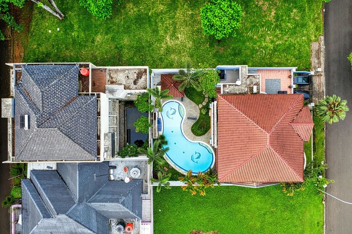 Aerial view of a home with a curved swimming pool, a red-tiled roof, and dark-grey gabled roofs surrounded by green lawns.