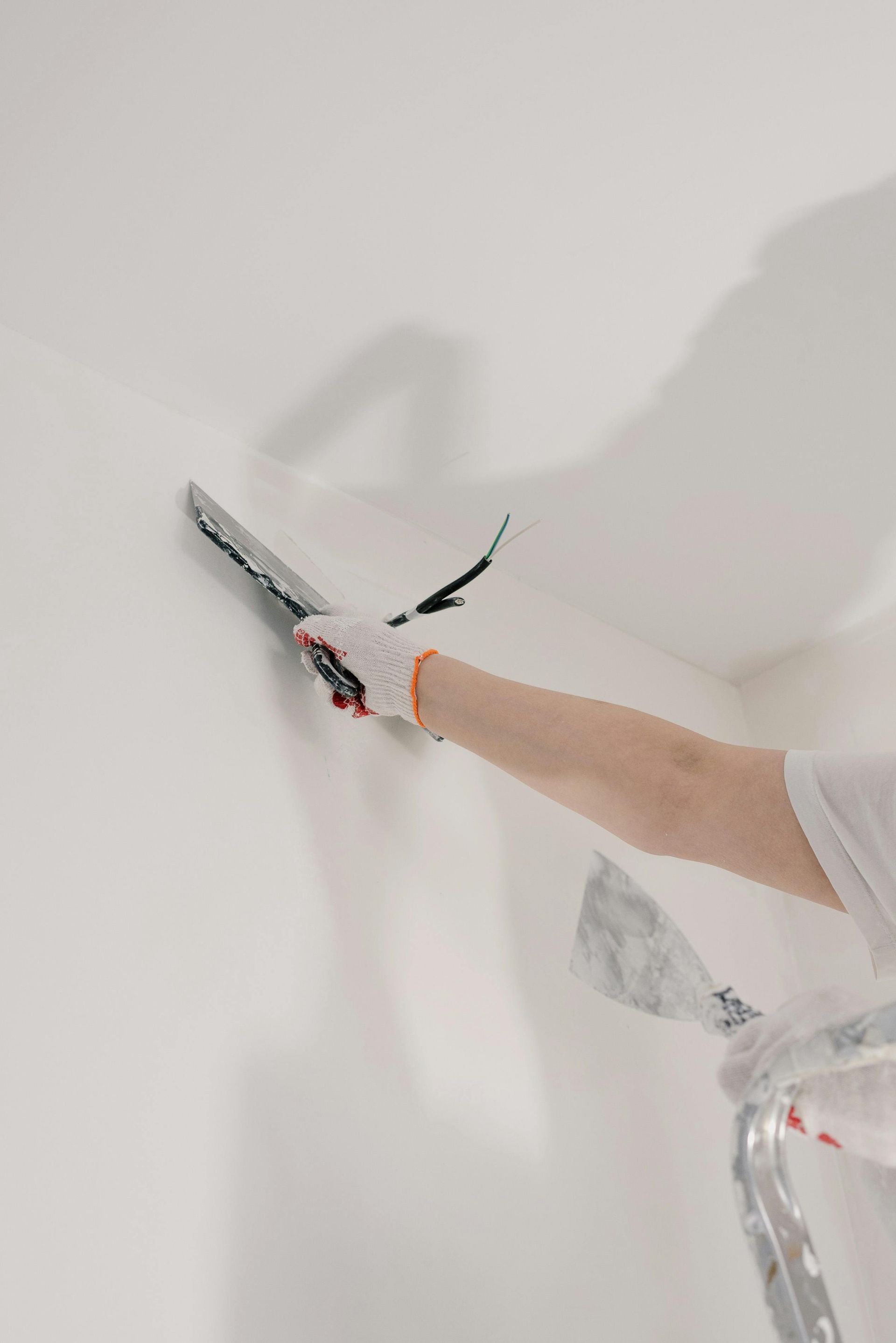 A person wearing a white glove uses a metal spatula to apply white plaster to a white interior wall.