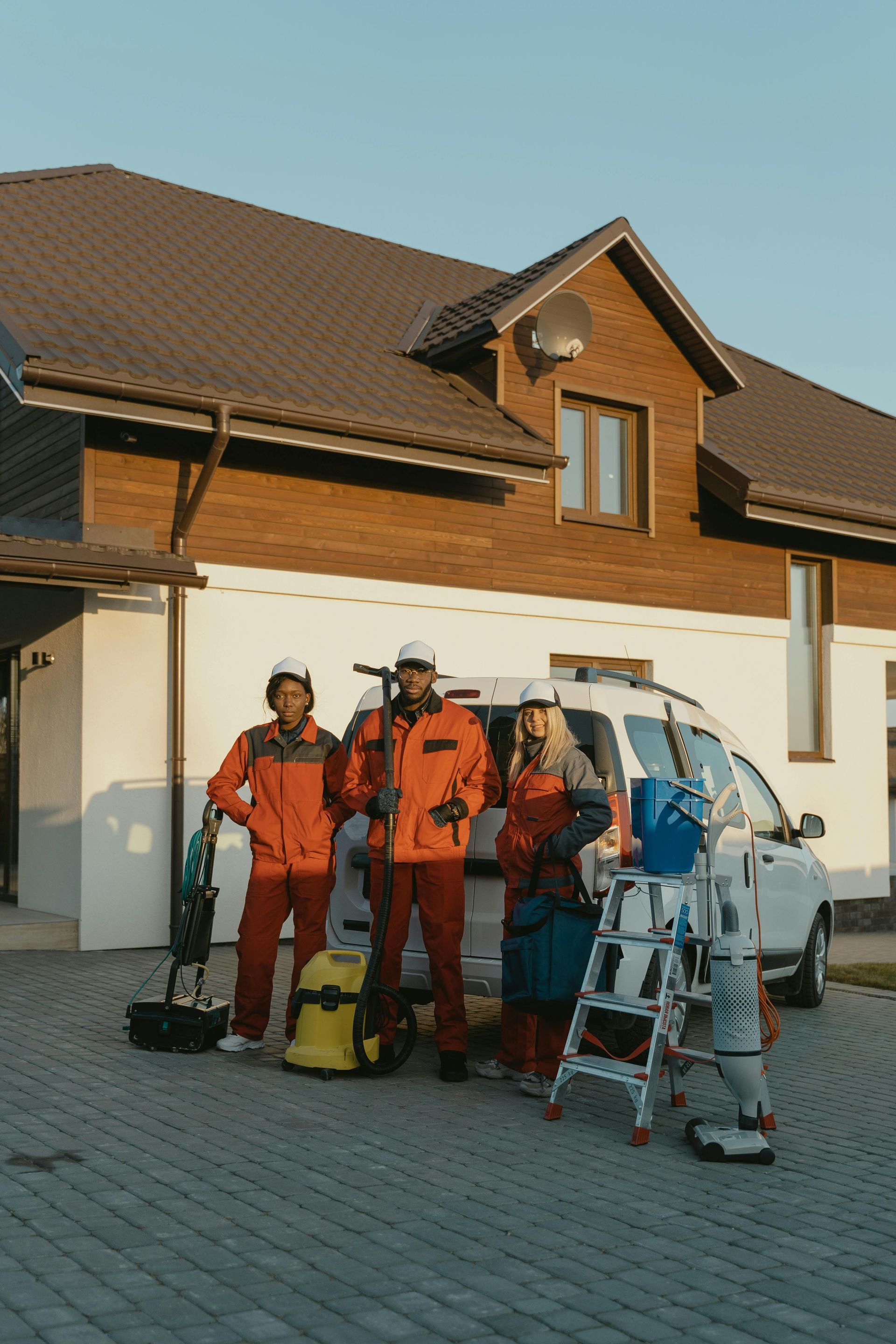 Three workers in orange uniforms stand with cleaning equipment and a van in front of a house.