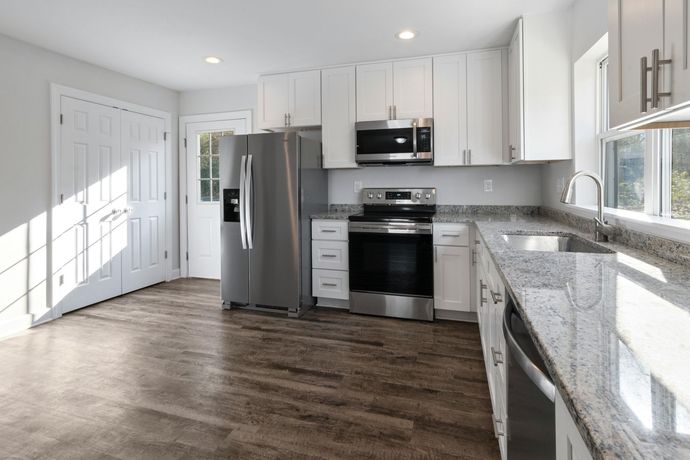 A bright, modern kitchen with white cabinets, stainless steel appliances, speckled granite countertops, and wood flooring.