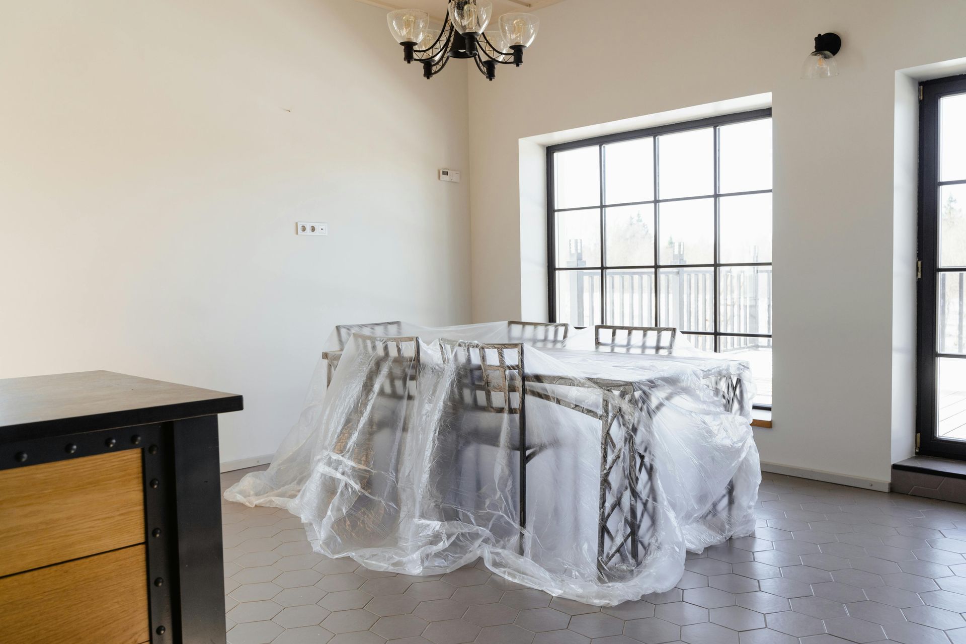 A dining table and chairs covered in clear protective plastic, situated in a minimalist room with large black-framed windows.
