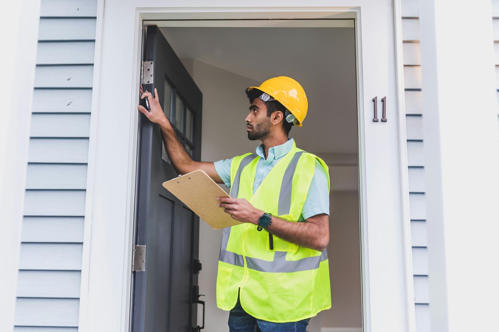 A person in a yellow hard hat and reflective vest inspects a door frame while holding a clipboard outside house number 11.