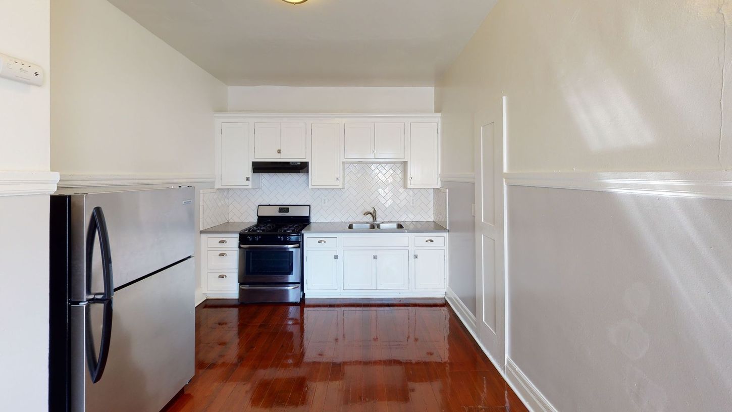 Empty, renovated kitchen with white cabinets, stainless steel refrigerator, and dark hardwood floor.