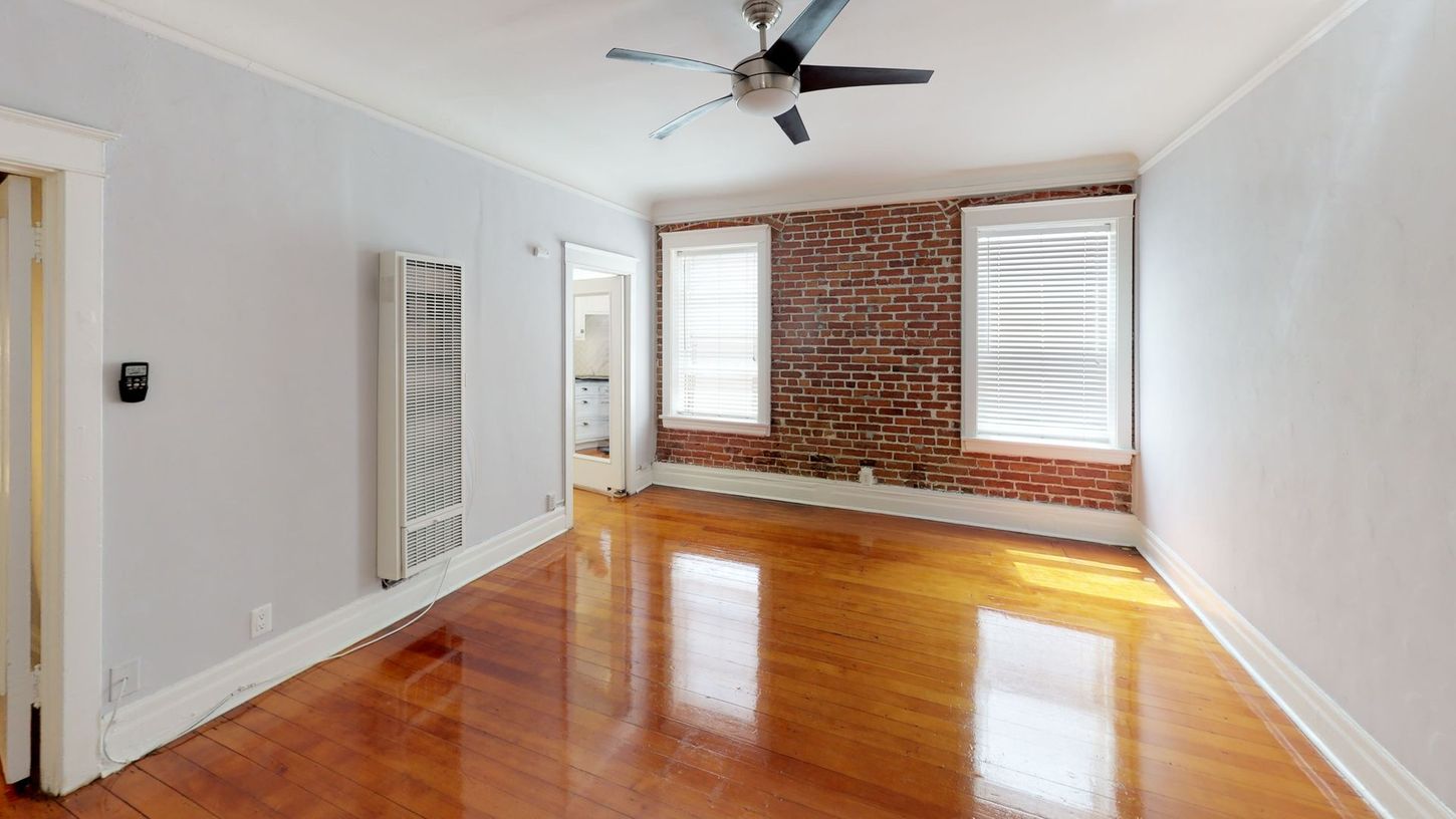 Empty room with hardwood floors, brick wall, two windows, radiator, and a ceiling fan.