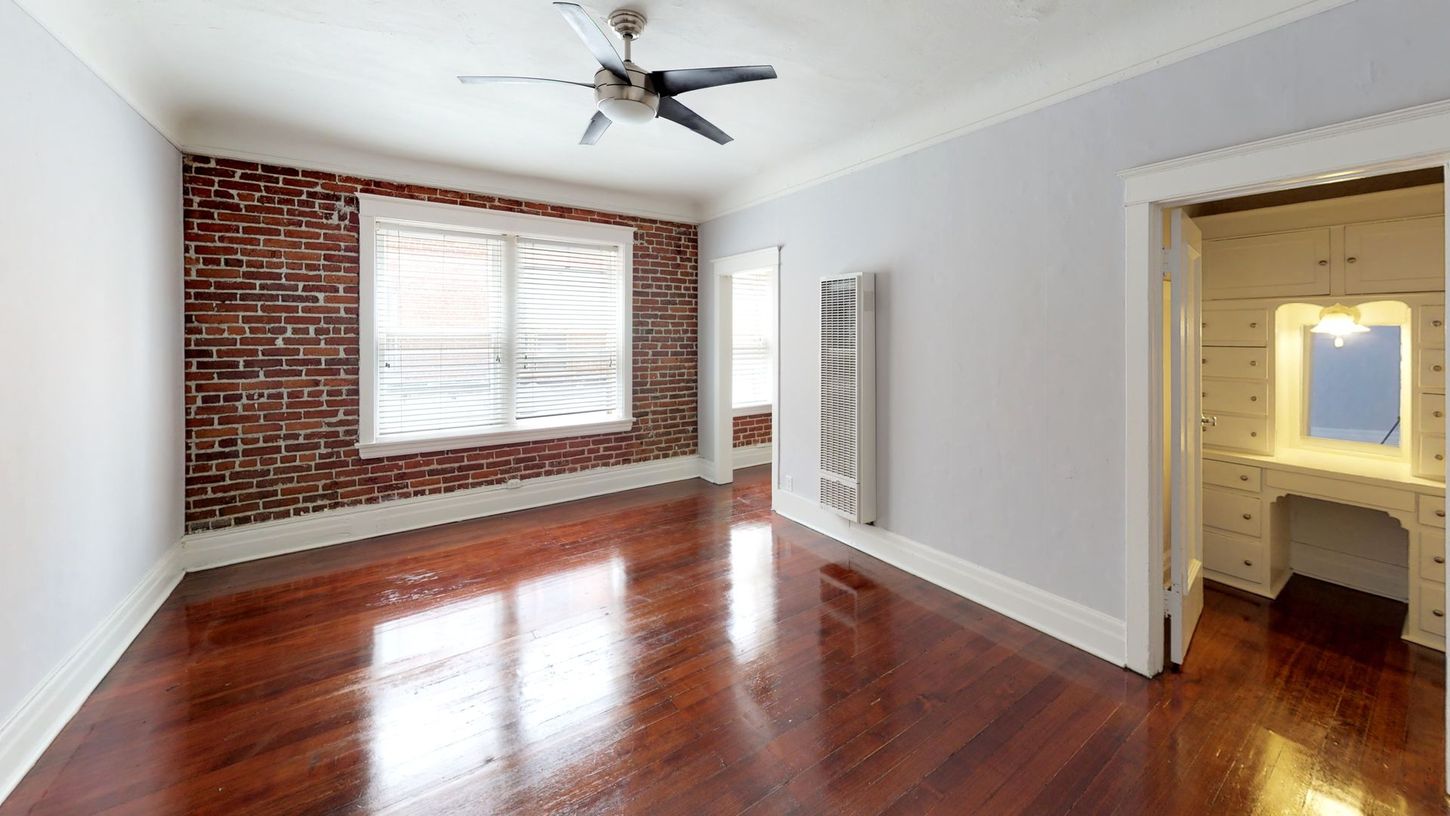 Empty room with exposed brick wall, wood floors, ceiling fan, and doorway to a vanity area.