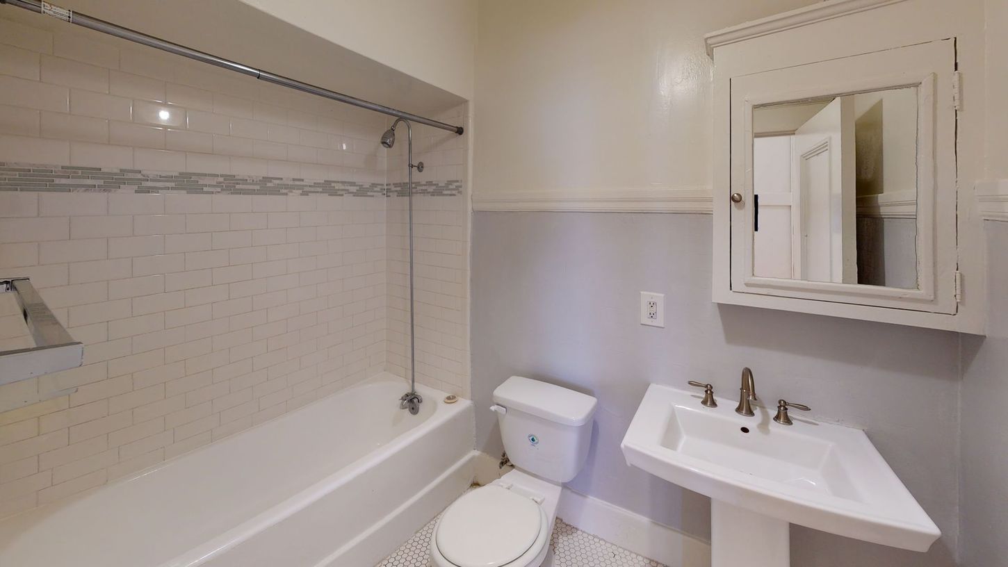Small, light-colored bathroom with a tub, toilet, pedestal sink, and medicine cabinet.