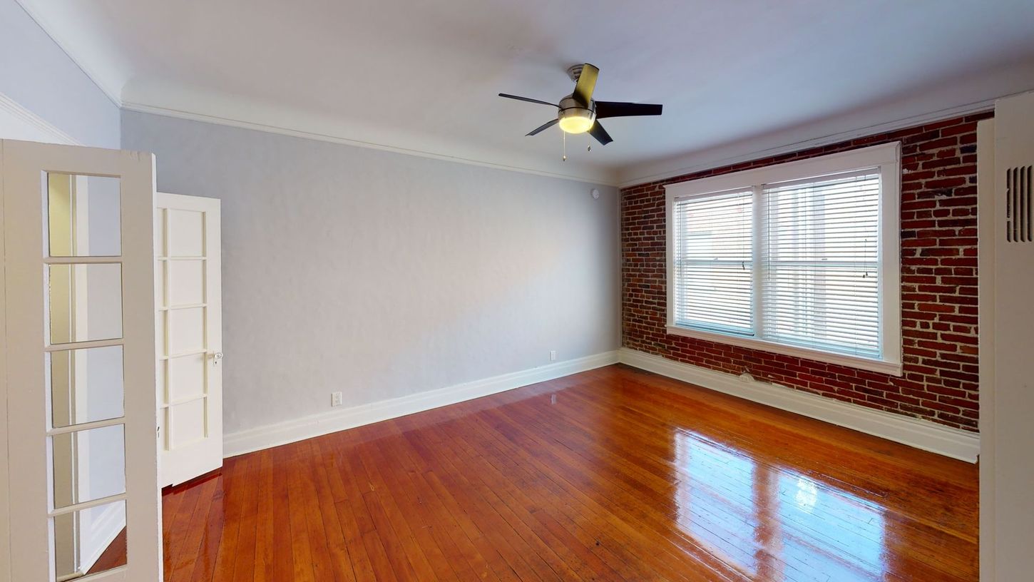 Empty room with hardwood floors, exposed brick wall, window, and white trim. Ceiling fan.