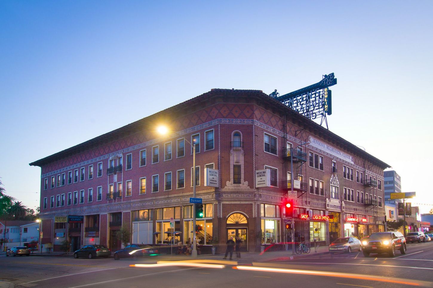 Brick building at a street corner with shops and traffic; a large illuminated sign is visible atop.