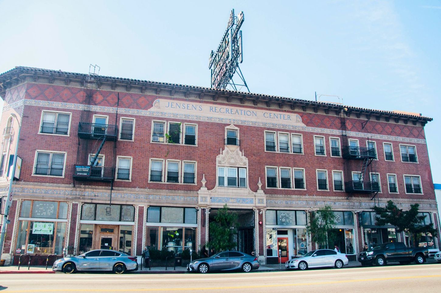 Red brick building with a sign on the roof, multiple windows, fire escapes, and cars parked in front.