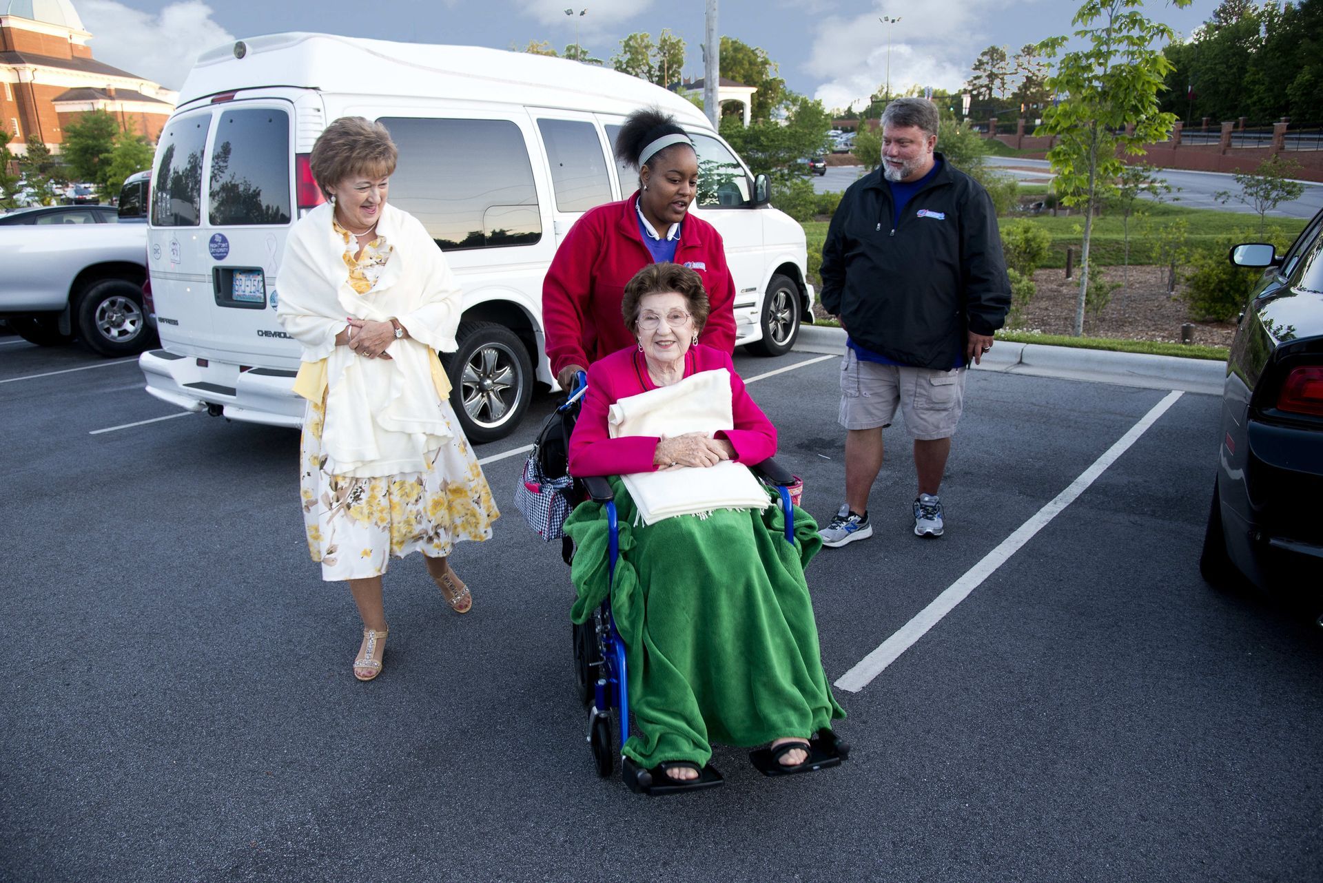 Person in wheelchair pushed by caregiver, others walking near a van.