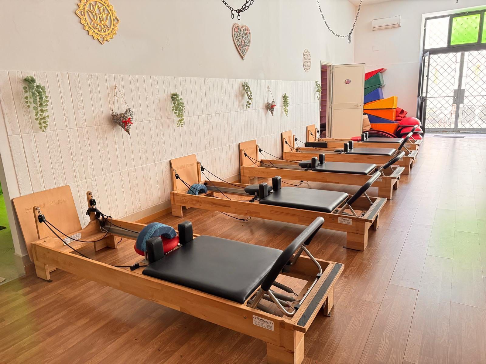 Studio di Pilates con reformer in legno, parete bianca testurizzata e ornamenti decorativi appesi.