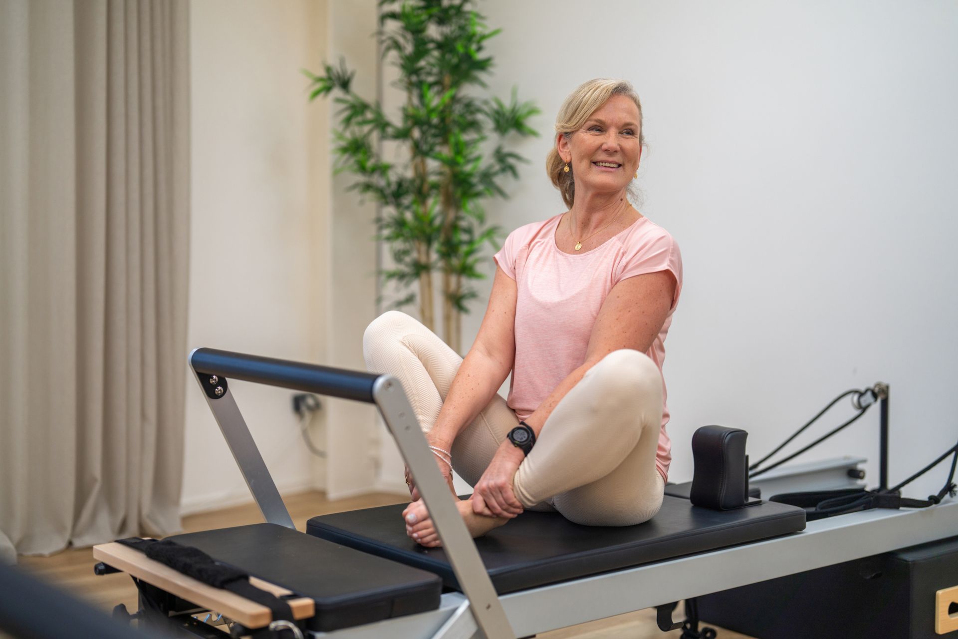 Una donna con top rosa e leggings sorride mentre è seduta su un reformer per Pilates in una stanza luminosa.
