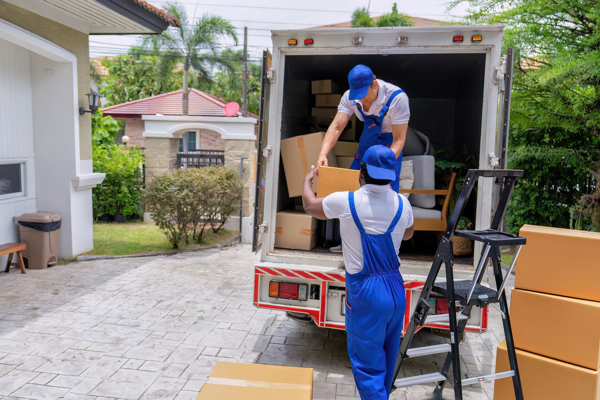 Two movers in blue coveralls loading boxes into a moving truck in front of a house.