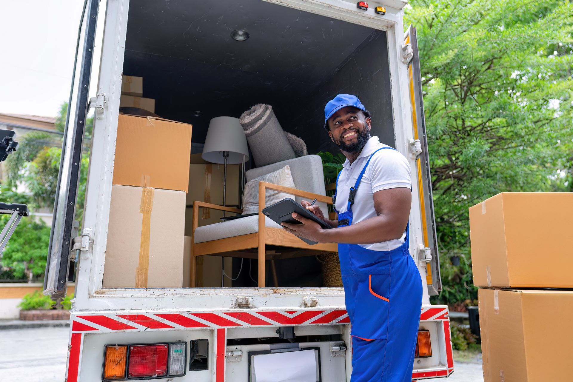 Moving worker with boxes in truck. Man in blue overalls and cap smiles, holding a tablet.