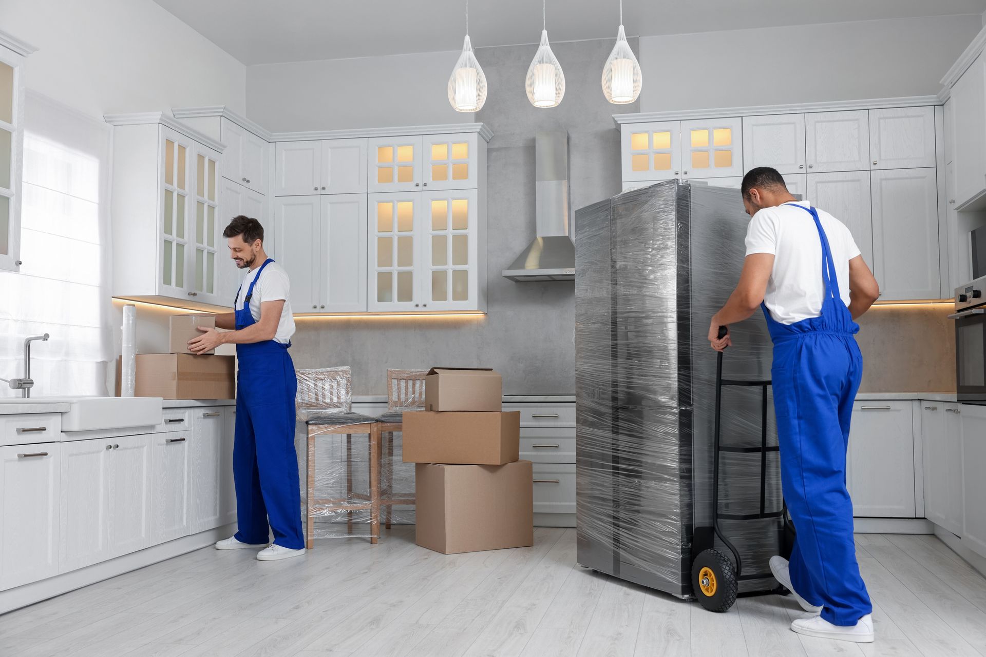 Two movers in blue overalls packing boxes and moving a wrapped refrigerator in a white kitchen.
