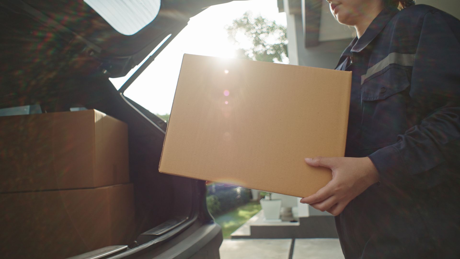 Person in blue uniform removes a cardboard box from the trunk of a car, sunny outdoors.