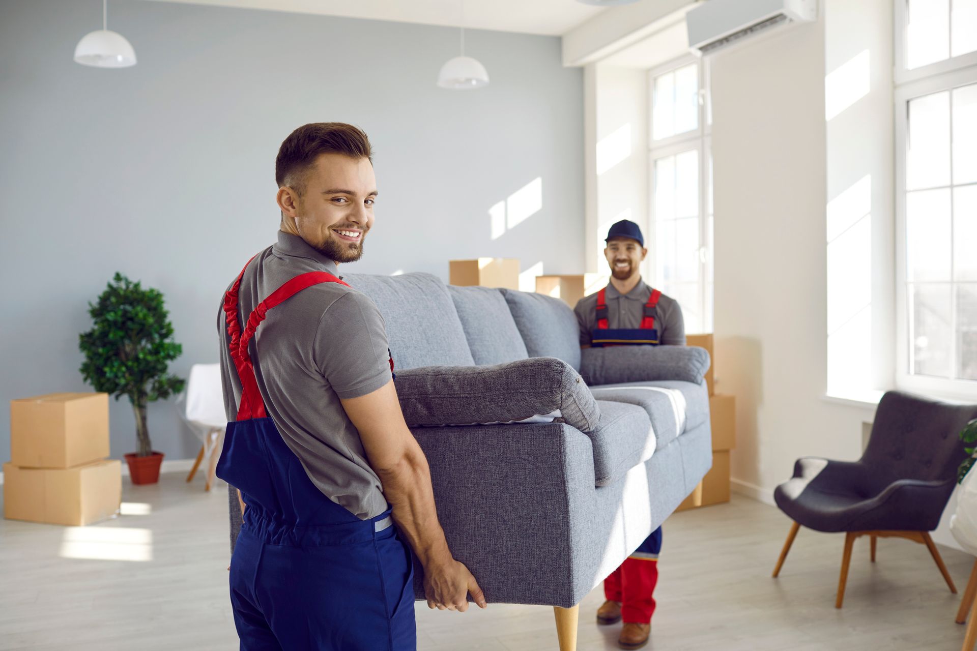 Two movers carrying a gray sofa in a room with boxes and furniture; one smiles.