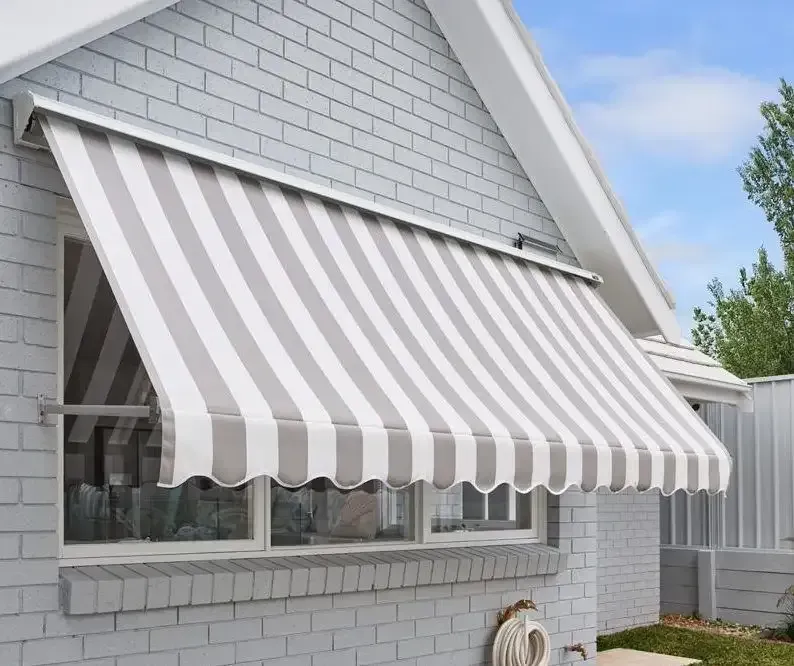 A White Brick House With a Striped Awning Over a Window — Elegant Blinds & Awnings in Forster, NSW
