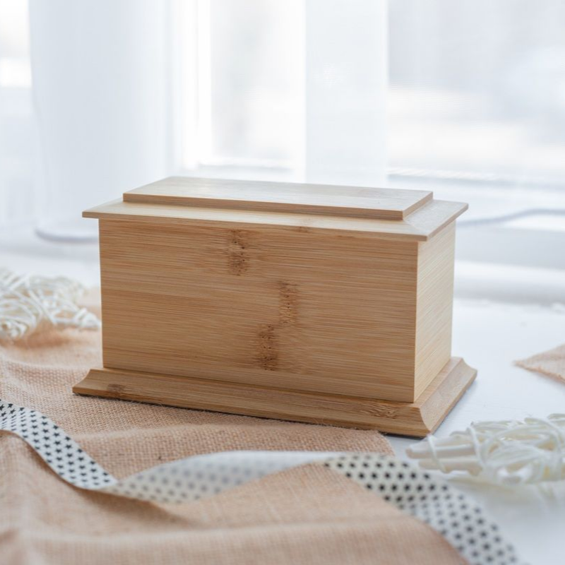 Wooden rectangular cremation urn with lid, on a burlap-like fabric with polka-dot ribbon, set before a window.
