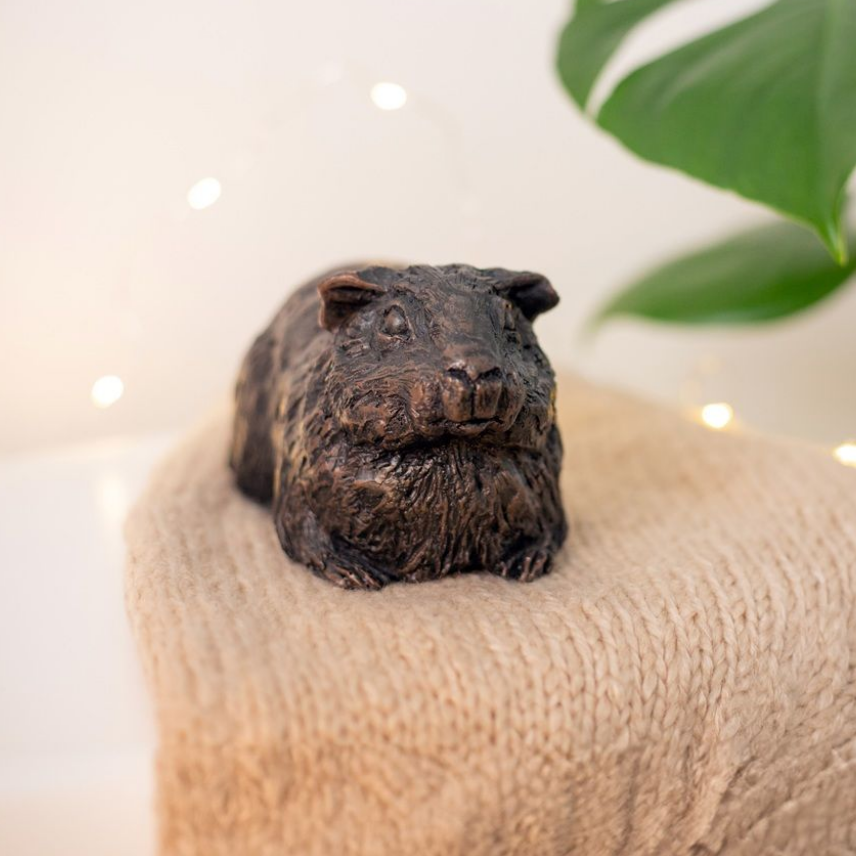 Bronze-colored guinea pig statue resting on a light brown knitted surface, with soft background bokeh.