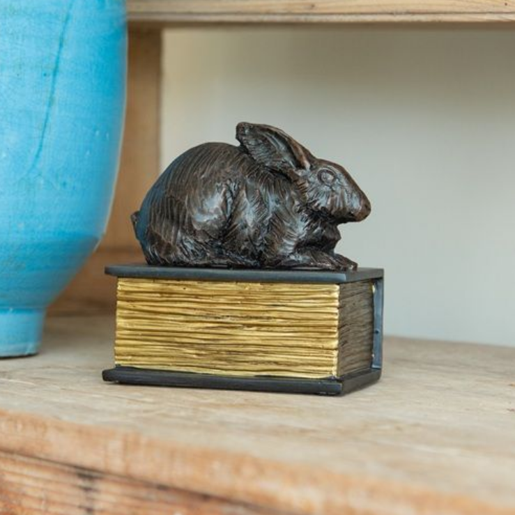 Bronze rabbit sculpture atop a gold-edged book on a wooden shelf, beside a blue vase.
