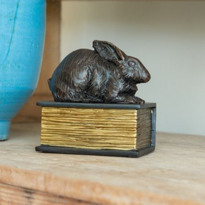 Bronze rabbit statue atop a small book with gold-colored pages, sitting on a wooden shelf near a blue vase.