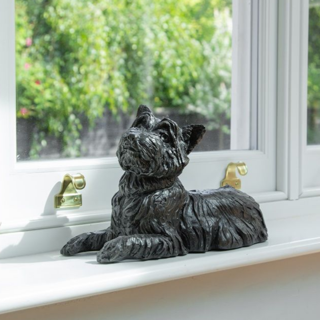 Black dog sculpture resting on a white windowsill, with a blurred green background.