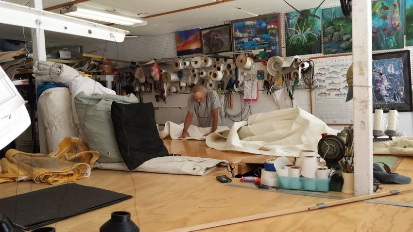 A person works on a large piece of white fabric atop a wooden workbench in a cluttered sail-making or upholstery studio.