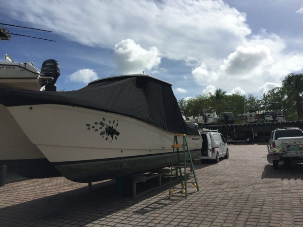 A white boat on a stand in a paved lot, covered with a black canopy, with a stepladder nearby under a partly cloudy sky.