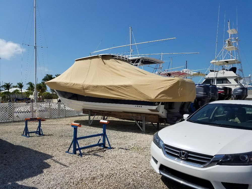 A white car is parked in a gravel lot next to a boat covered by a tan tarp on supports under a bright blue sky.