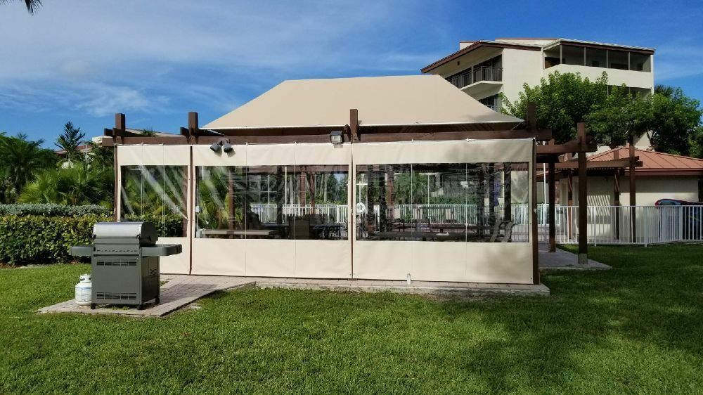 A covered outdoor pavilion with clear plastic siding, set in a grassy yard next to a barbecue grill and a large building.