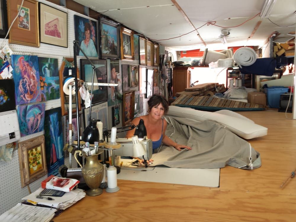 A person works on a large piece of beige fabric with a sewing machine in a room decorated with many framed paintings.