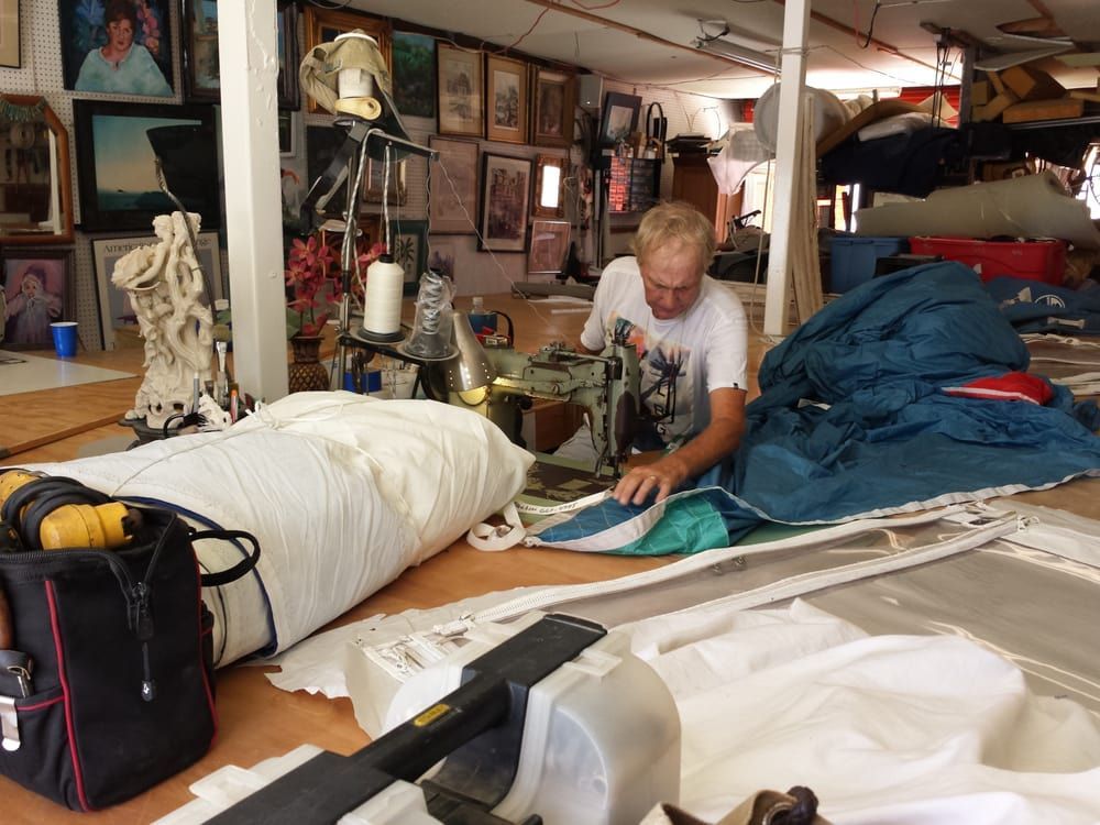 A person in a white t-shirt operates a heavy-duty sewing machine, repairing a large blue fabric panel in a workshop.