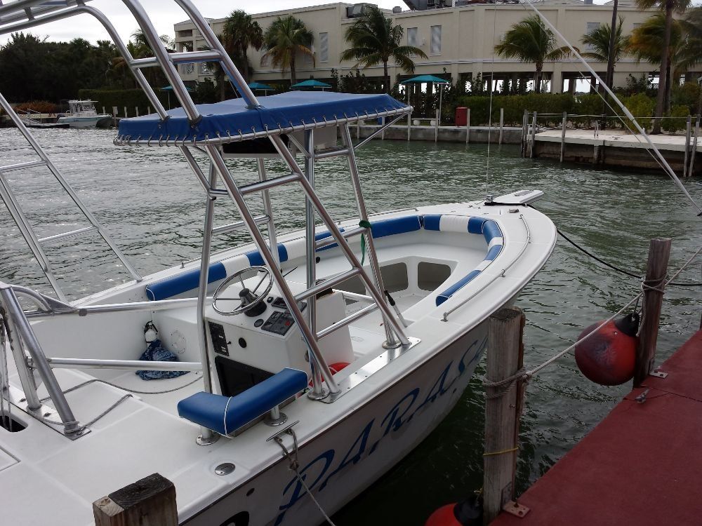 A white center-console boat with a blue canopy and cushions docked at a pier near tropical buildings.