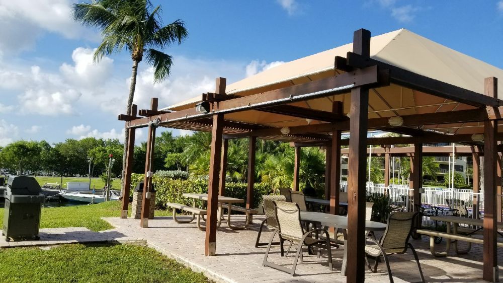 An outdoor, wood-framed pavilion with a canopy, patio furniture, and a grill near a waterway and greenery.