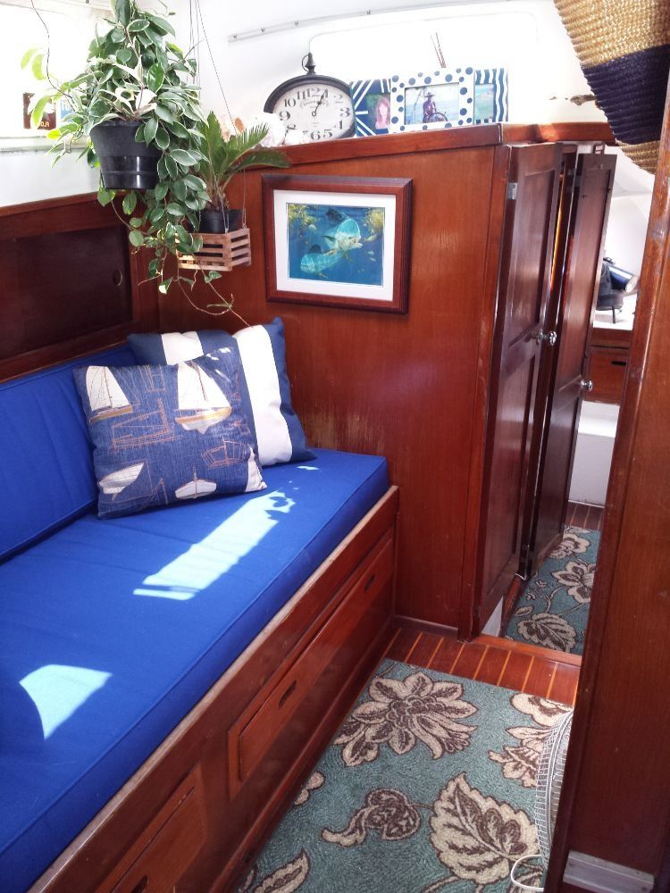 Interior of a boat cabin with a blue cushioned bench, wooden walls, a framed picture, hanging plants, and a floral rug.