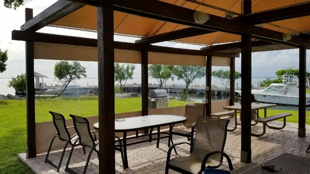 A covered outdoor patio with tables and chairs overlooking a green lawn, a marina, and a boat.