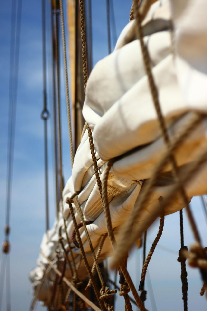 A close-up of a tightly furled white sail on a wooden ship mast, with ropes and rigging against a clear blue sky.