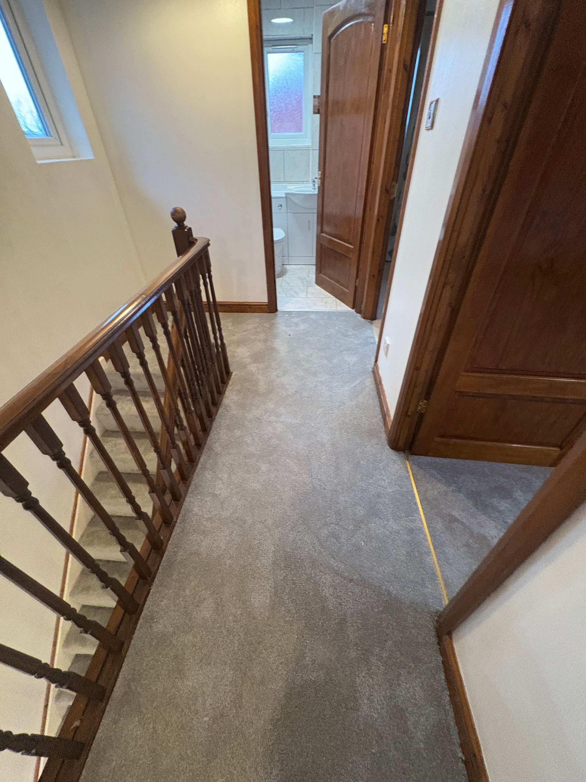 Hallway with gray carpet, wood doors and banister, leading to a bathroom.