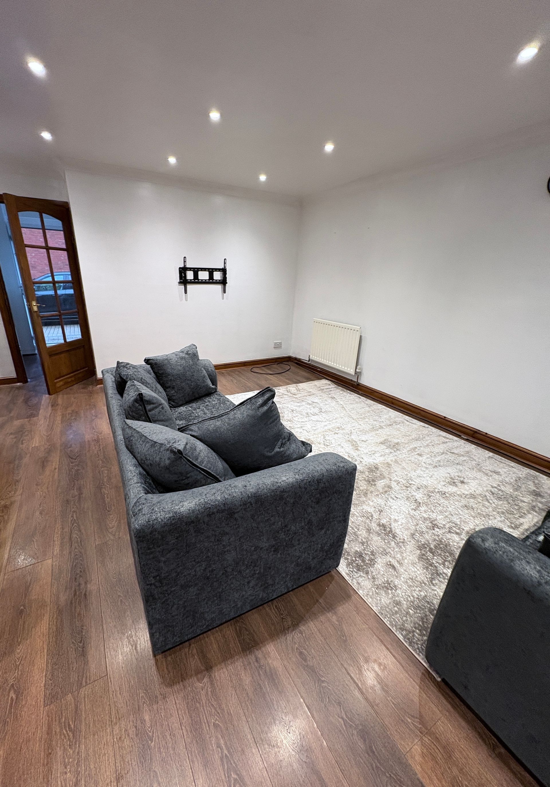 Gray sofa on a rug, mounted TV bracket on the wall, door on the left, wood floor.