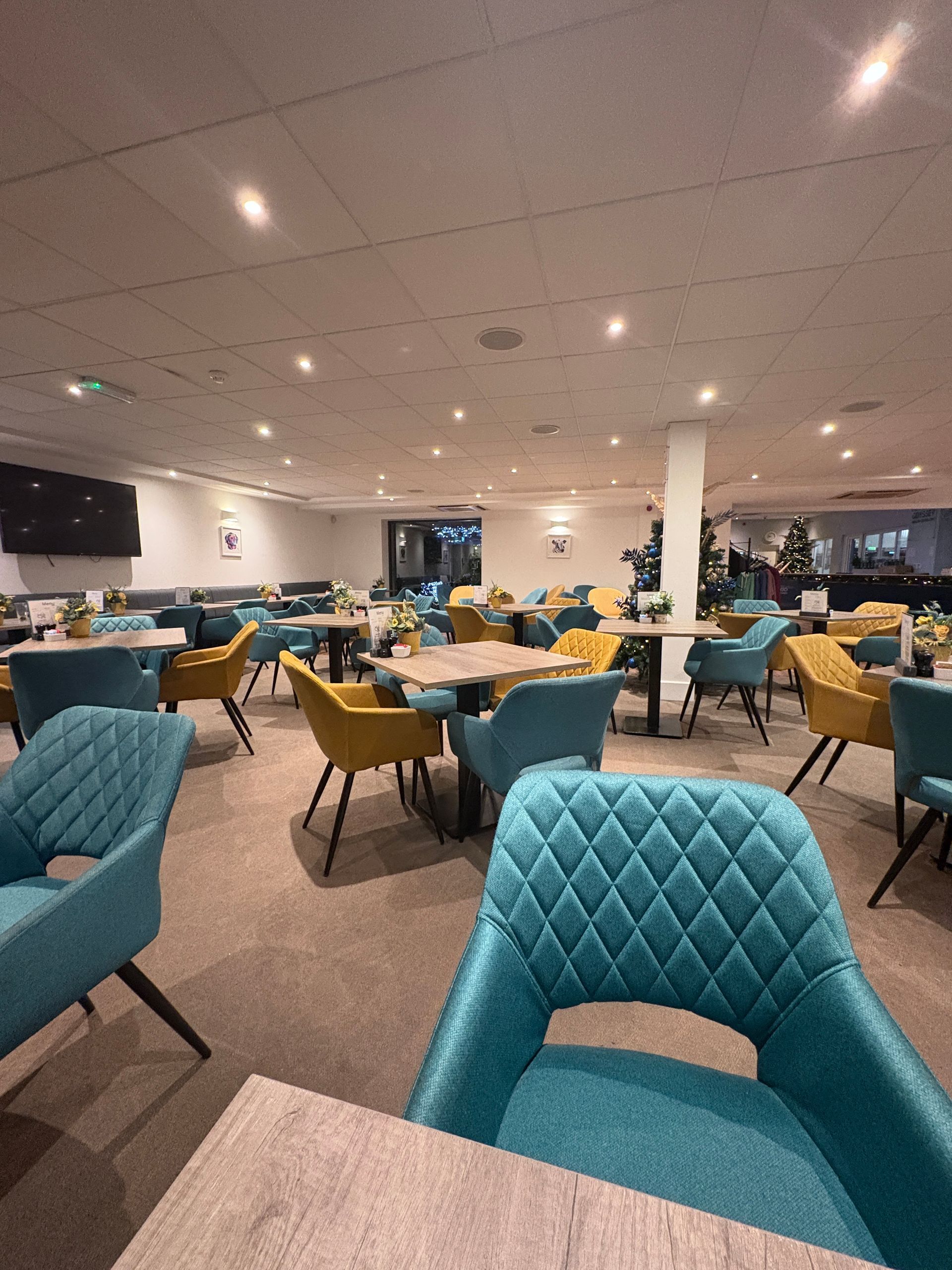 Interior view of a modern restaurant with teal and yellow chairs around tables. Beige carpet and ceiling.